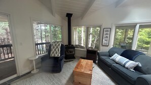 A living room with a fireplace, a sofa, a wooden coffee table, and a view of trees through large windows.