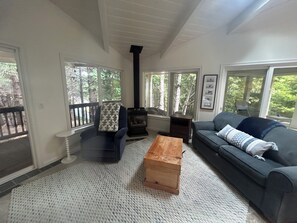 A living room with a fireplace, a sofa, a wooden coffee table, and a view of trees through large windows.