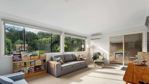Un salón amplio con un sofá gris, una estantería con libros infantiles, una mesa de centro de madera y grandes ventanales con vistas a zonas verdes.