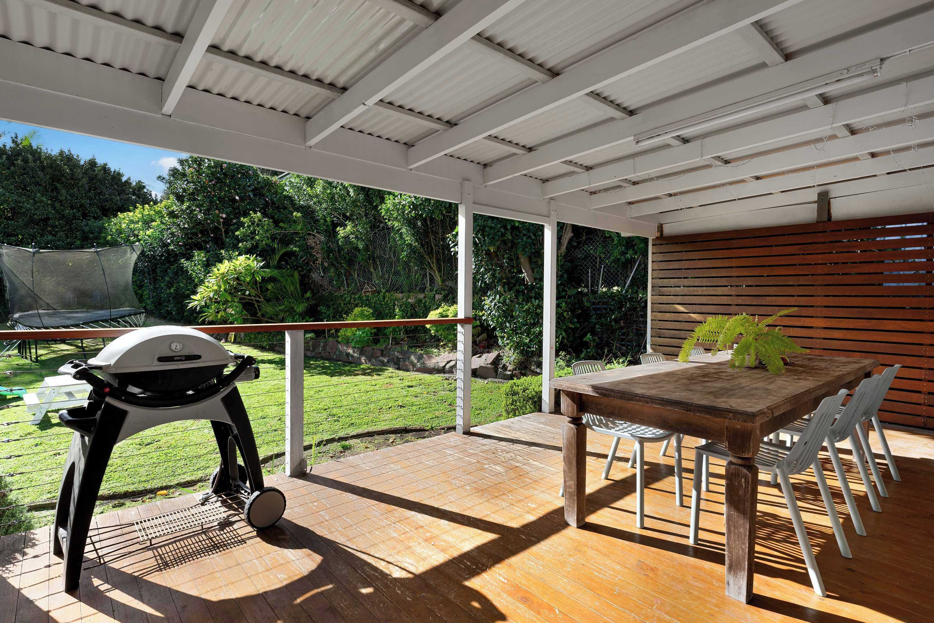 An outdoor patio with a wooden table and chairs, a grill, and a view of a lush garden.