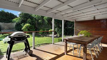 An outdoor patio with a wooden table and chairs, a grill, and a view of a lush garden.