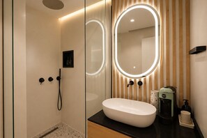 Une salle de bain moderne avec une douche à parois de verre, un meuble-lavabo doté d’un grand miroir et une machine à café posée sur le plan de travail.