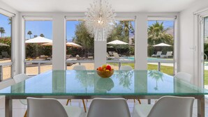 A modern dining area with a glass table, white chairs, a fruit bowl, and a chandelier. - Mountain Mirage | MCM 3BR | Private Pool & Spa | Bocce | Sleeps 6 (Palm Springs)