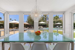 A modern dining area with a glass table, white chairs, a fruit bowl, and a chandelier. - Mountain Mirage | MCM 3BR | Private Pool & Spa | Bocce | Sleeps 6 (Palm Springs)