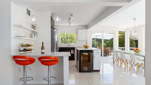 A modern kitchen with white cabinetry, a central island with bar stools, and a dining area with a glass table and chairs. - Mountain Mirage | MCM 3BR | Private Pool & Spa | Bocce | Sleeps 6 (Palm Springs)
