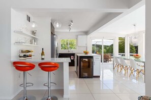 A modern kitchen with white cabinetry, a central island with bar stools, and a dining area with a glass table and chairs. - Mountain Mirage | MCM 3BR | Private Pool & Spa | Bocce | Sleeps 6 (Palm Springs)