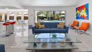 A modern living room with a glass coffee table, a blue sofa, and orange chairs. - Mountain Mirage | MCM 3BR | Private Pool & Spa | Bocce | Sleeps 6 (Palm Springs)