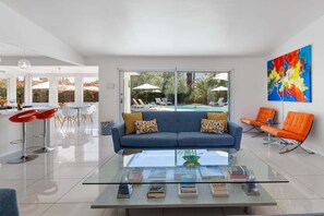 A modern living room with a glass coffee table, a blue sofa, and orange chairs. - Mountain Mirage | MCM 3BR | Private Pool & Spa | Bocce | Sleeps 6 (Palm Springs)