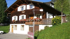 A traditional wooden chalet with a balcony adorned with red flowers, a sign indicating a restaurant, and a view of a lush green hillside.