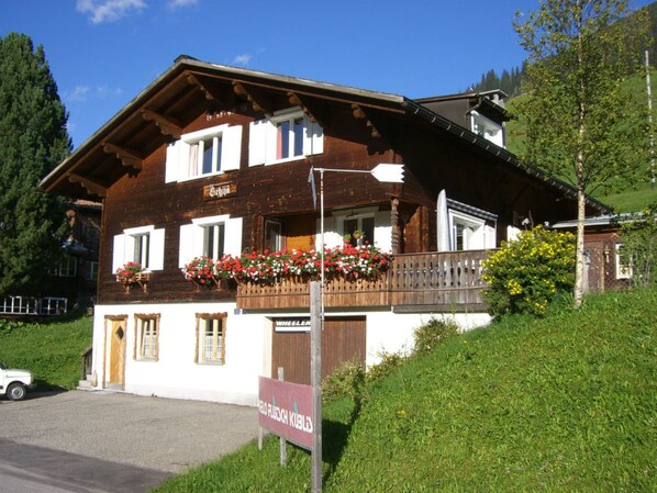 A traditional wooden chalet with a balcony adorned with red flowers, a sign indicating a restaurant, and a view of a lush green hillside.