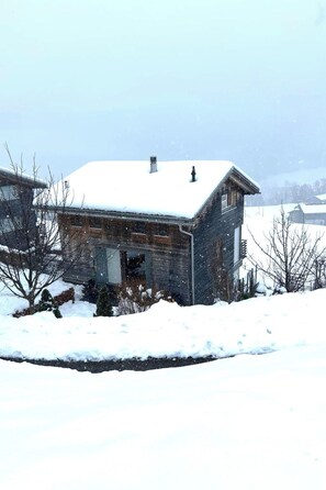 A wooden cabin with a snow-covered roof, surrounded by snow-covered ground and bare trees. - Ferienhaus Jukale, (Luzein), ferienhaus_jukale by Interhome (Luzein)