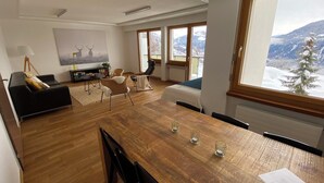 A modern living room with a wooden dining table, a sofa, and a view of a snowy mountain landscape through large windows. - Ferienwohnungen JHS, (Seewis), by Interhome (Seewis im Prättigau)