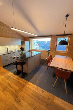 Una cocina moderna con una isla central, un comedor con mesa y sillas de madera, y vistas a montañas a través de una ventana.