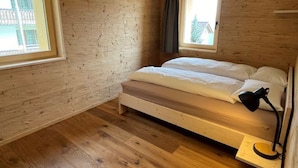 Makuuhuone, jossa on puinen, kalteva kattopinta, valkoisilla vuodevaatteilla varustettu sänky, yöpöytä ja ikkuna, josta on näkymä rakennuksiin.