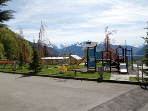 Un parque infantil con tobogán y estructuras para escalar, rodeado de árboles y con una montaña al fondo.