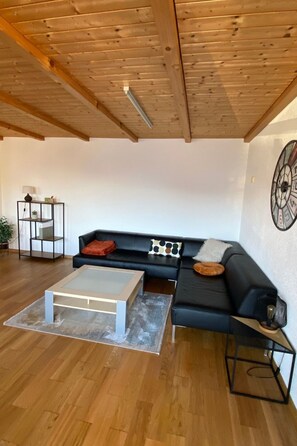 Una sala de estar moderna con techo de madera, un sofá negro con cojines, una mesa de centro de vidrio y una mesita auxiliar.