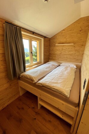 A wooden room with a bed, a window with curtains, and a sloped ceiling.
