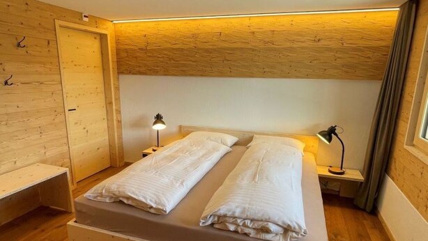 Una stanza con un letto di legno, due comodini, una panca e un soffitto spiovente.