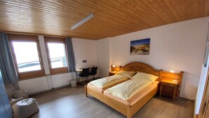 Ett sovrum med en säng i trä, två sängbord med lampor, ett skrivbord med en stol, ett element och ett fönster med gardiner.