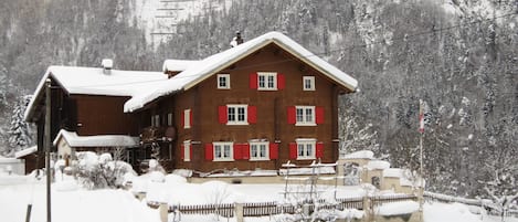 En stuga av trä med många fönster och ett sluttande tak, omgiven av snötäckta träd och en bergig bakgrund.