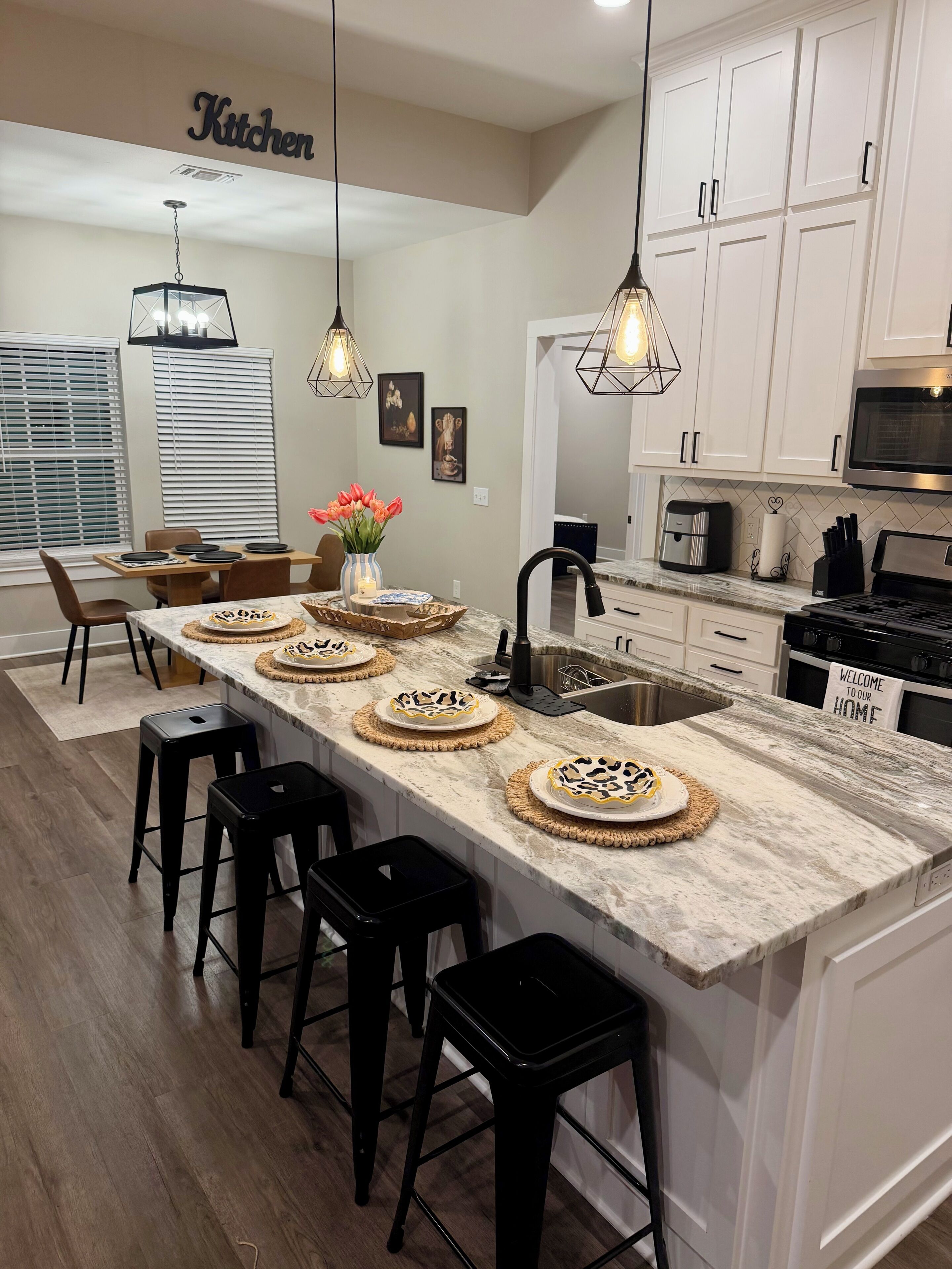 A modern kitchen with white cabinets, a marble island with bar stools, pendant lighting, and a 'Welcome to our home' sign.