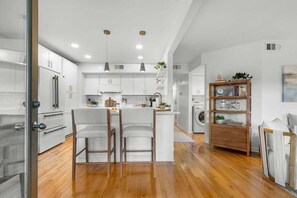 A modern kitchen with white cabinets, a central island with bar stools, pendant lighting, and a wooden sideboard with books and plants. - Charming 2BR/2BA Courtside Villa, near Equestrian Center and Tennis. (Seabrook Island)