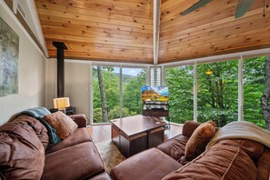 A cozy living room with a brown leather couch, a wooden ceiling, and a large window offering a view of greenery outside. - Cozy High Country Retreat Near the Slopes (Sugar Mountain)