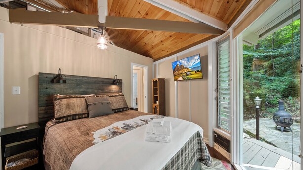 A bedroom with a wooden ceiling, a bed with a floral bedspread, a nightstand, a wall-mounted lamp, and a window with a view of greenery. - Cozy High Country Retreat Near the Slopes (Sugar Mountain)