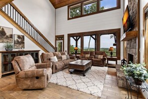 A spacious living room with a fireplace, large windows, and a high ceiling. - Talona Rising Sun - Mountain Views | Pet Friendly | Firepit | Game Room (Ellijay)