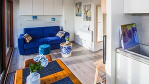 Ein modernes Wohnzimmer mit einem blauen Sofa, einem kleinen runden Tisch mit einer Pflanze und einer Küchenzeile mit Kühlschrank und Spüle.