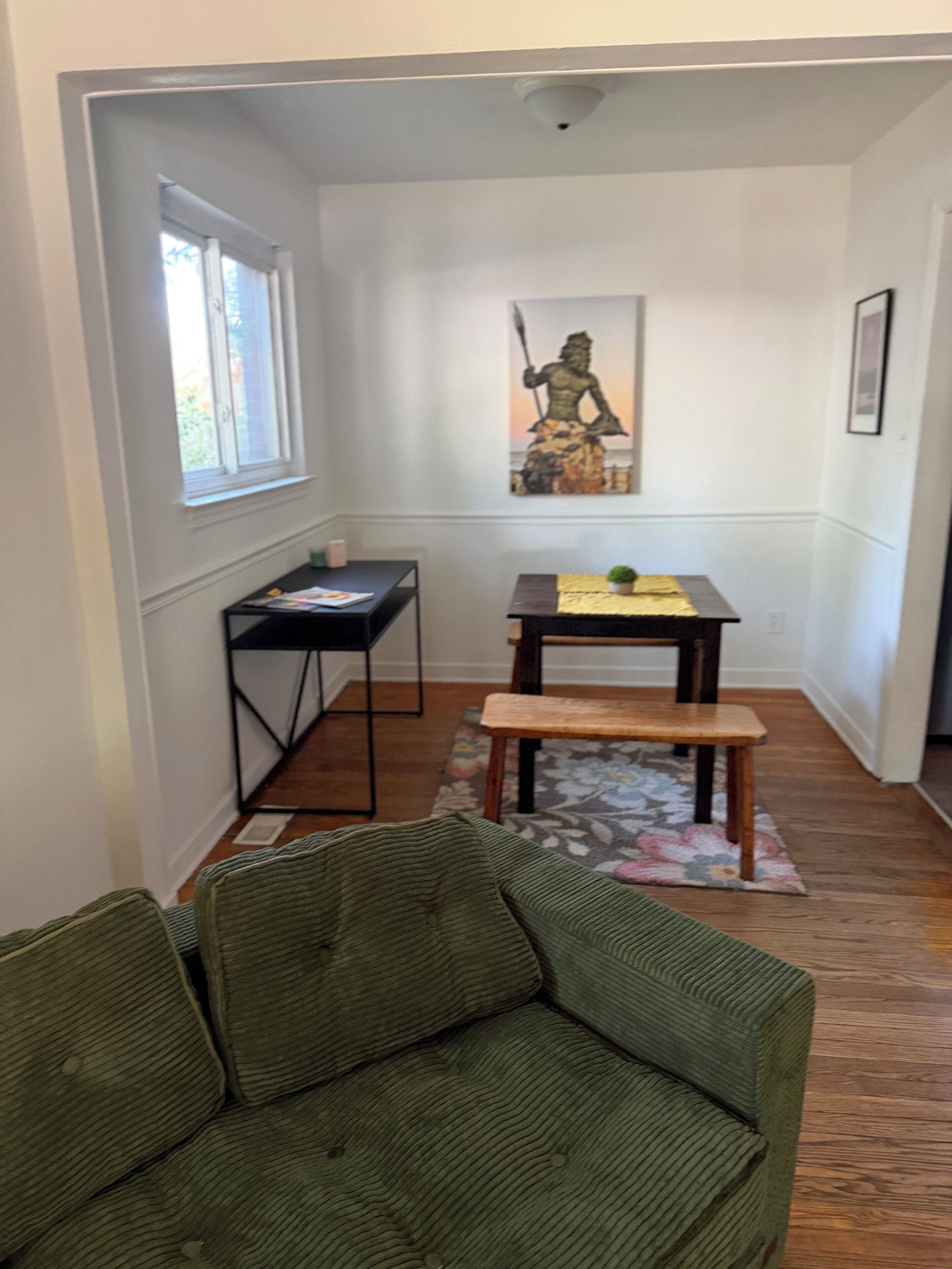 A room with a green sofa, a wooden dining table with a bench, a small potted plant, and a statue on the wall.