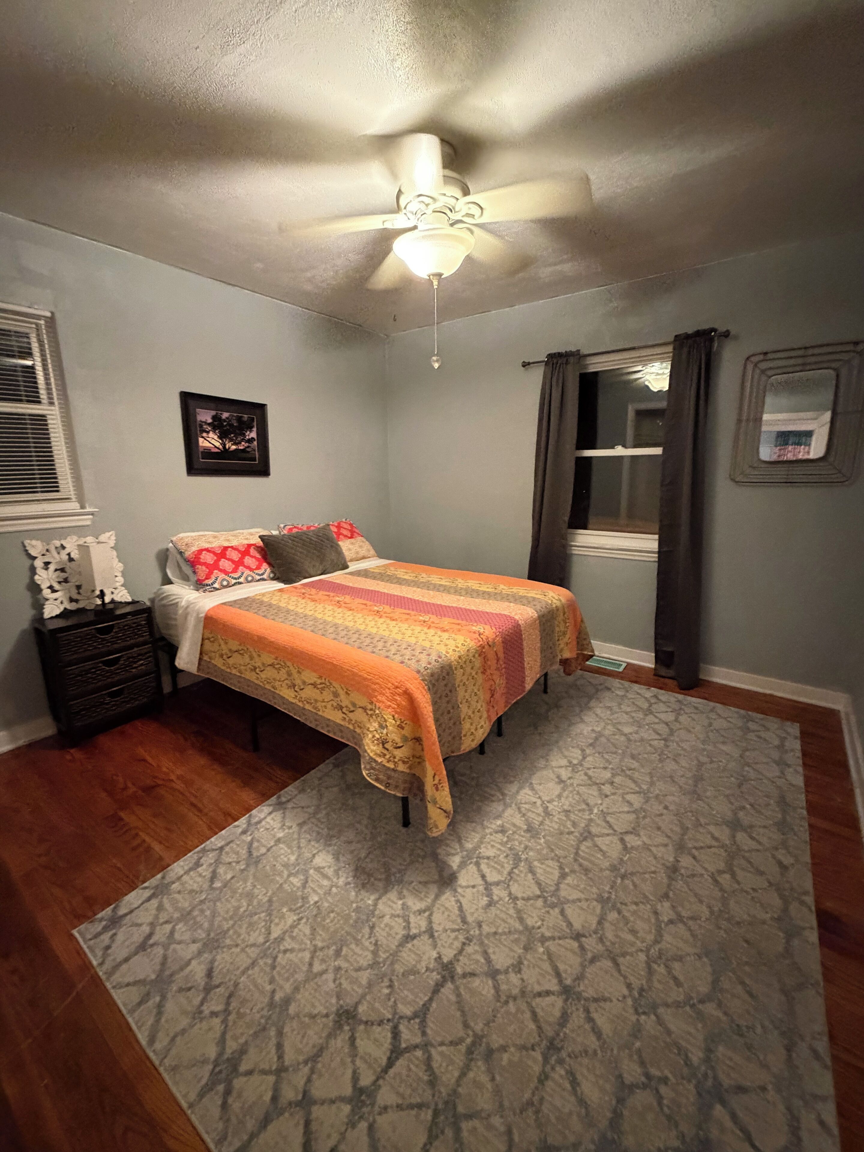 A bedroom with a bed, a ceiling fan, a window with curtains, a framed picture, and a small dresser.