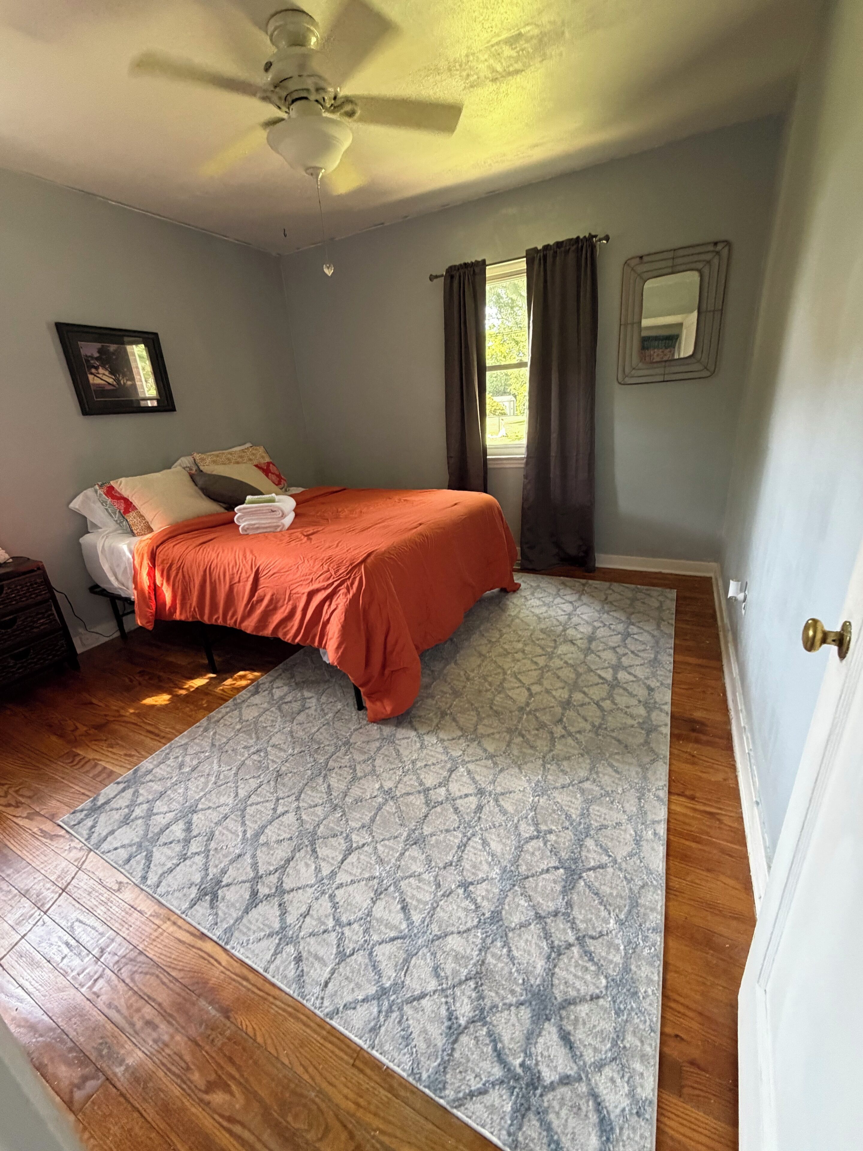 A bedroom with a bed, a ceiling fan, a window with curtains, and a patterned rug.