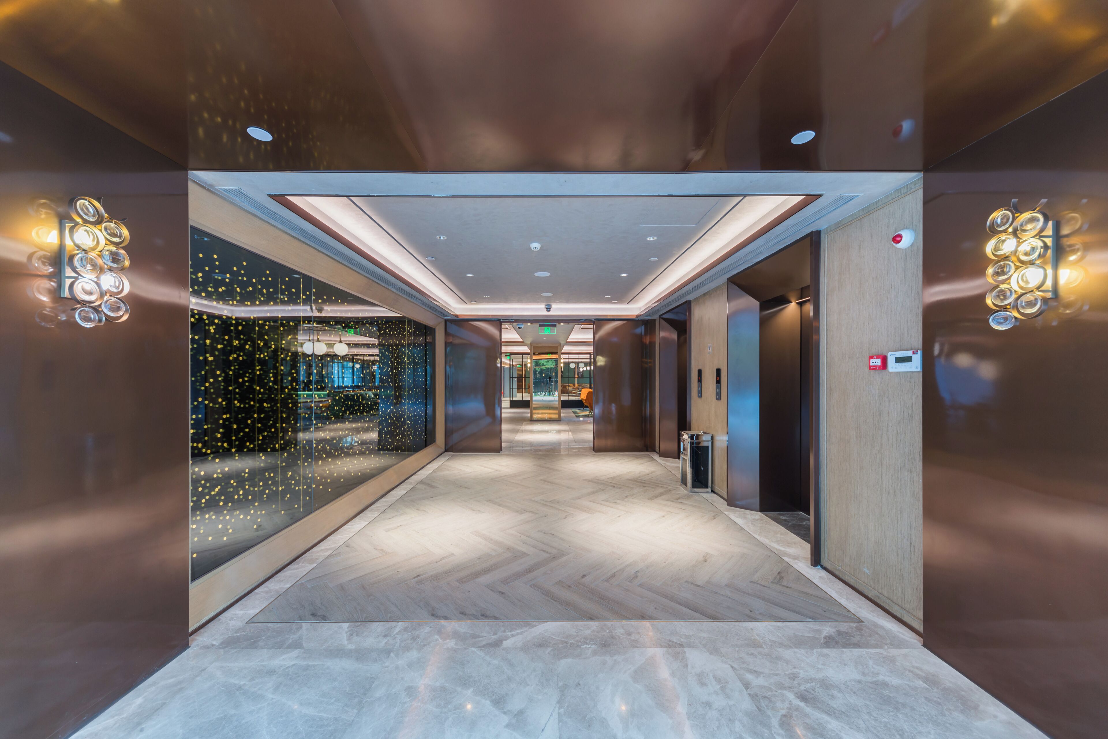 A modern elevator interior with a herringbone floor pattern, recessed lighting, and a glass wall with decorative lights.