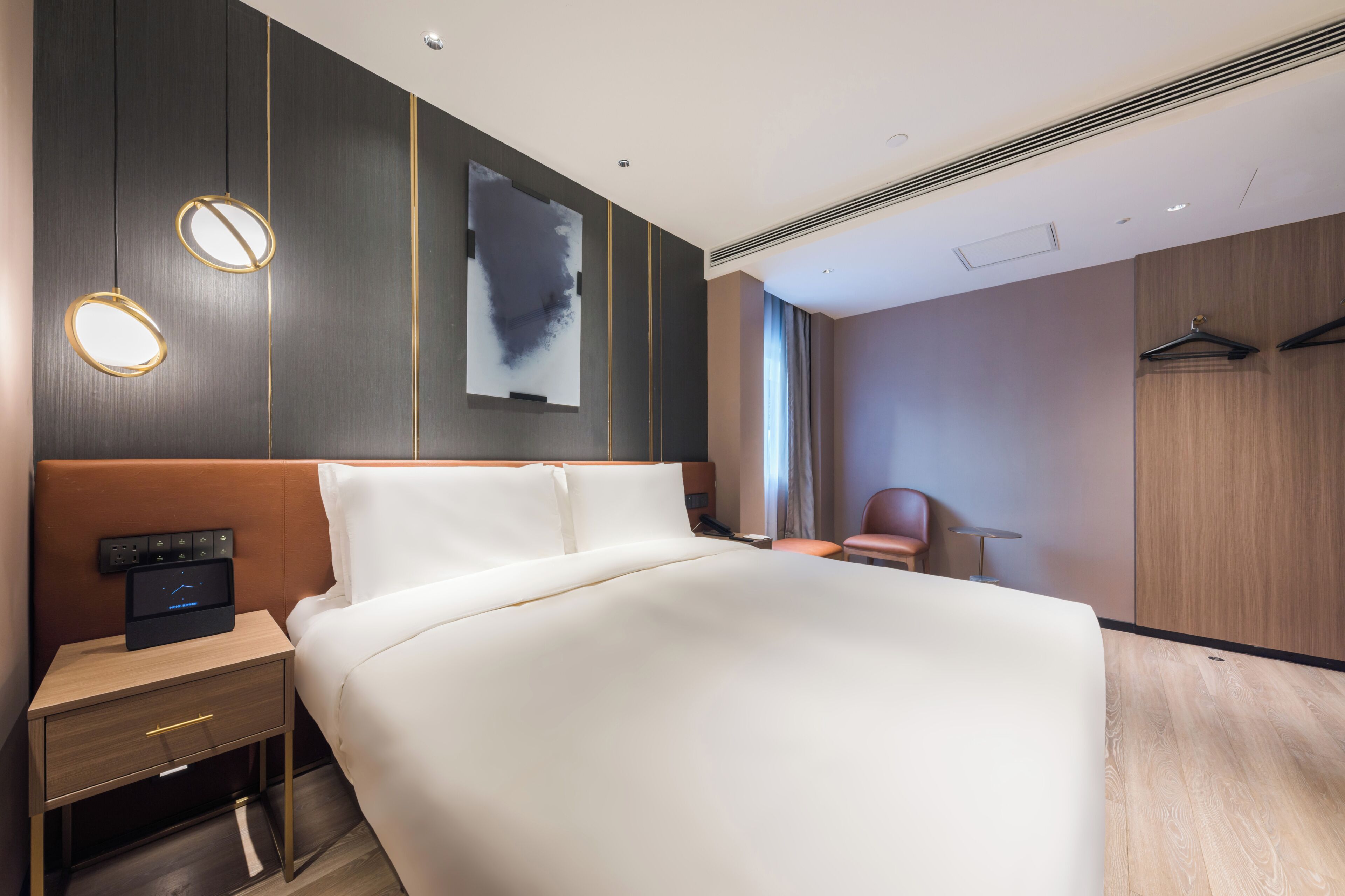 A modern hotel room with a large bed, bedside table with a control panel, a chair, and a wardrobe.