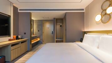 A modern hotel room with a large bed, a vanity with a mirror, a shower area, and a small seating area with a table.