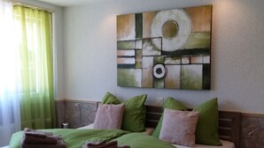 Hotelkamer met een bed, groene en roze kussens, een kunstwerk aan de muur en een raam met dunne gordijnen.
