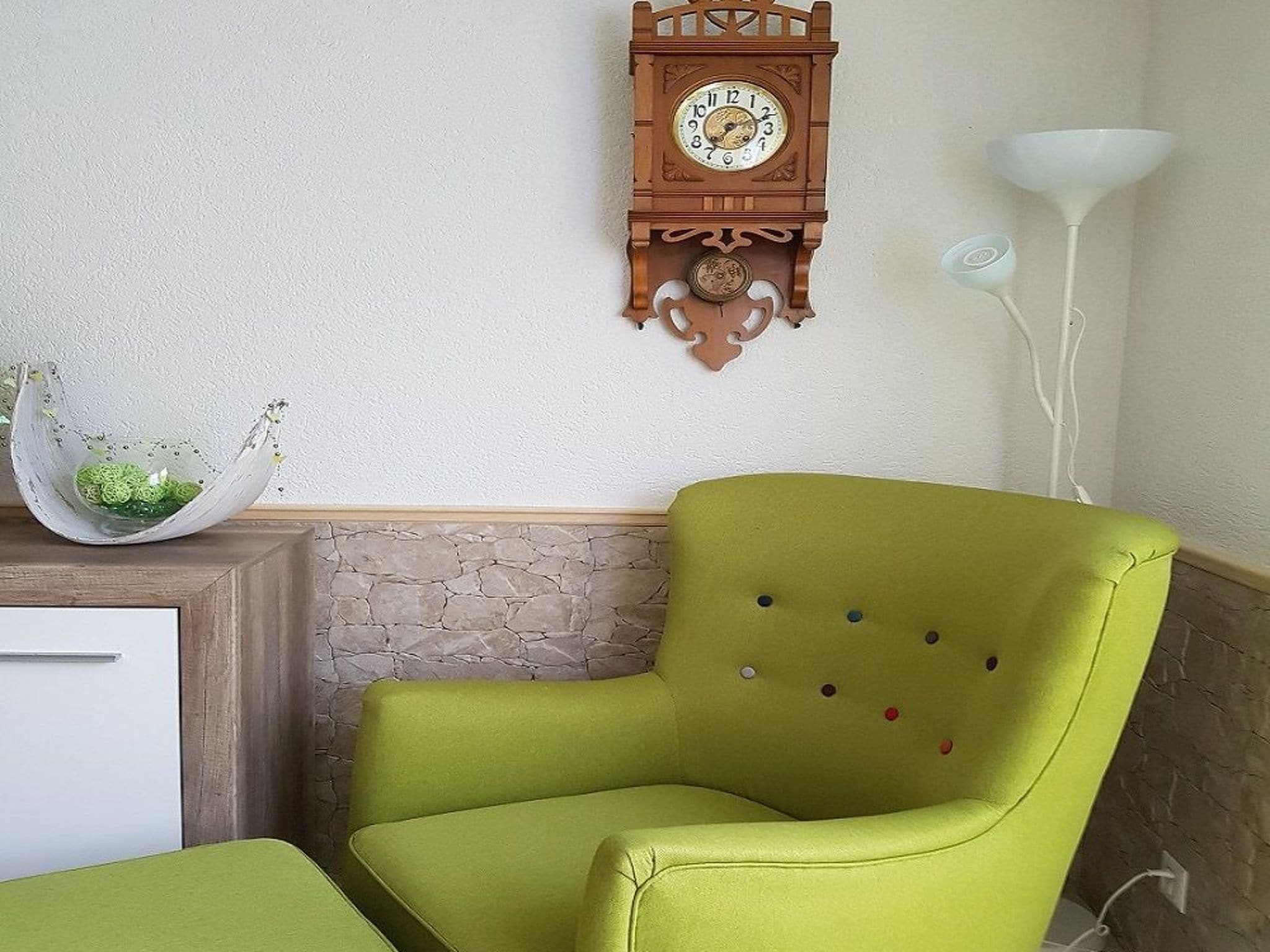 A room with a green armchair, a wooden clock, a white lamp, and a stone wall.