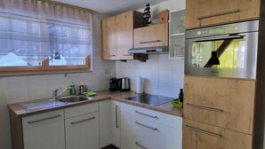 Een kleine keuken met witte kastjes, een roestvrijstalen spoelbak, een inbouwoven en een raam met transparante gordijnen.