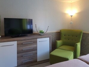 Habitación de hotel con televisor sobre un mueble de madera, un sillón verde y una mesita de noche.
