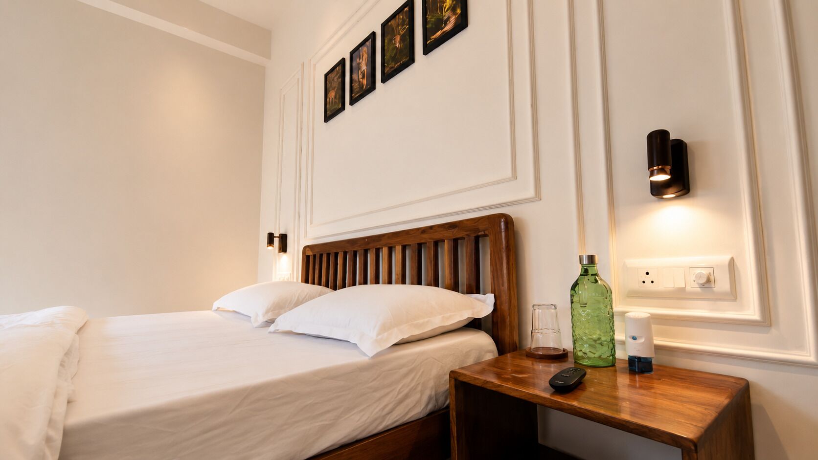 Habitación de hotel con cama de madera, ropa de cama blanca, mesita de noche con un control remoto, una botella de vidrio y un aplique de pared.