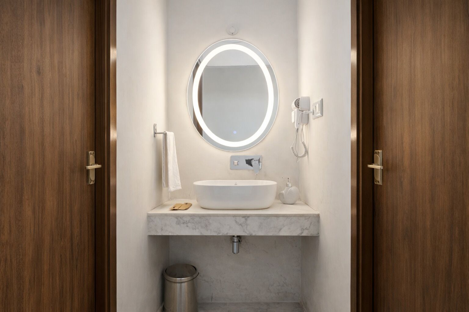 Un baño con espejo redondo, lavamanos blanco, toalla, secador de pelo y un cesto de basura.