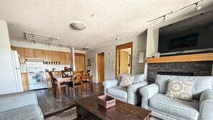 Ein Wohnzimmer mit Sofa, Couchtisch und Fernseher. Es gibt eine Küchenzeile mit Holzschränken und einem Herd.