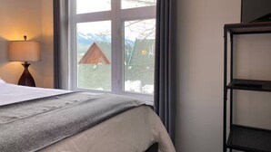 Ein Hotelzimmer mit Bett, Lampe, Fenster mit Blick auf ein Haus und Berge sowie einem schwarzen Metallregal.