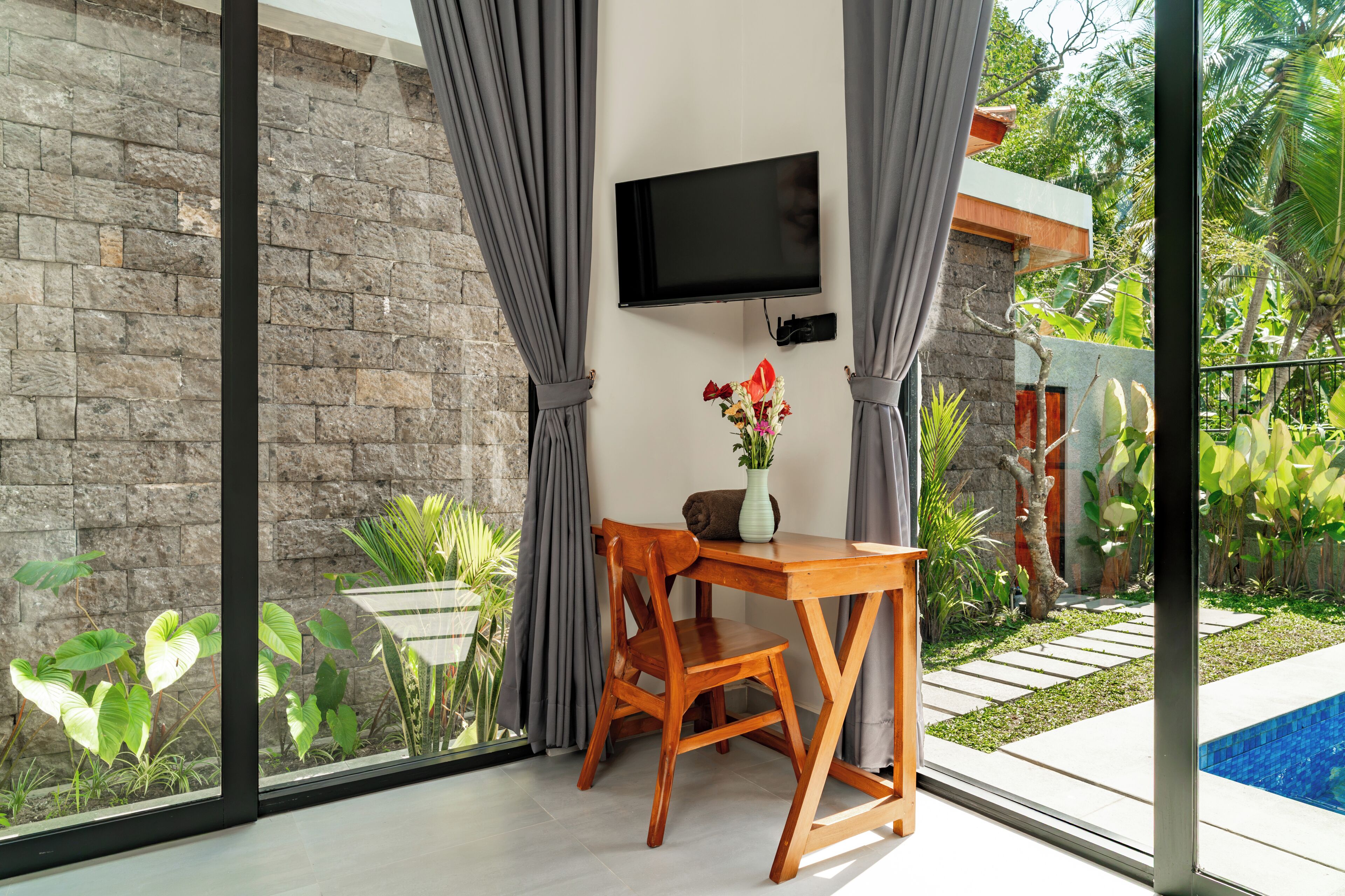 A room with a stone wall, a flat-screen TV, a wooden table with a chair, a vase with flowers, and a view of a swimming pool and greenery.