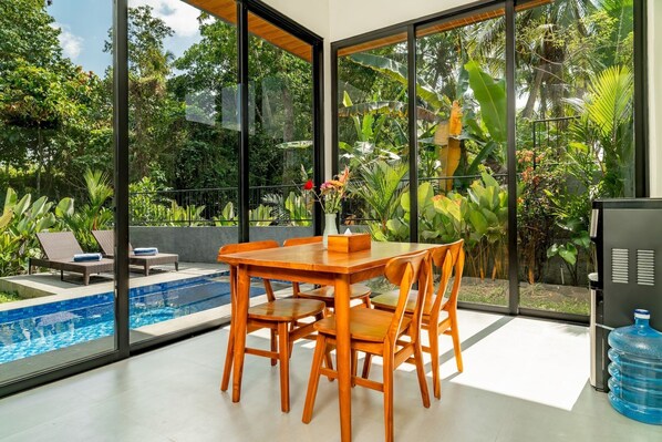 A modern indoor space with a pool, wooden furniture, and large windows offering a view of lush greenery. - Bugenvil Villa Ubud (Kabupaten Gianyar)