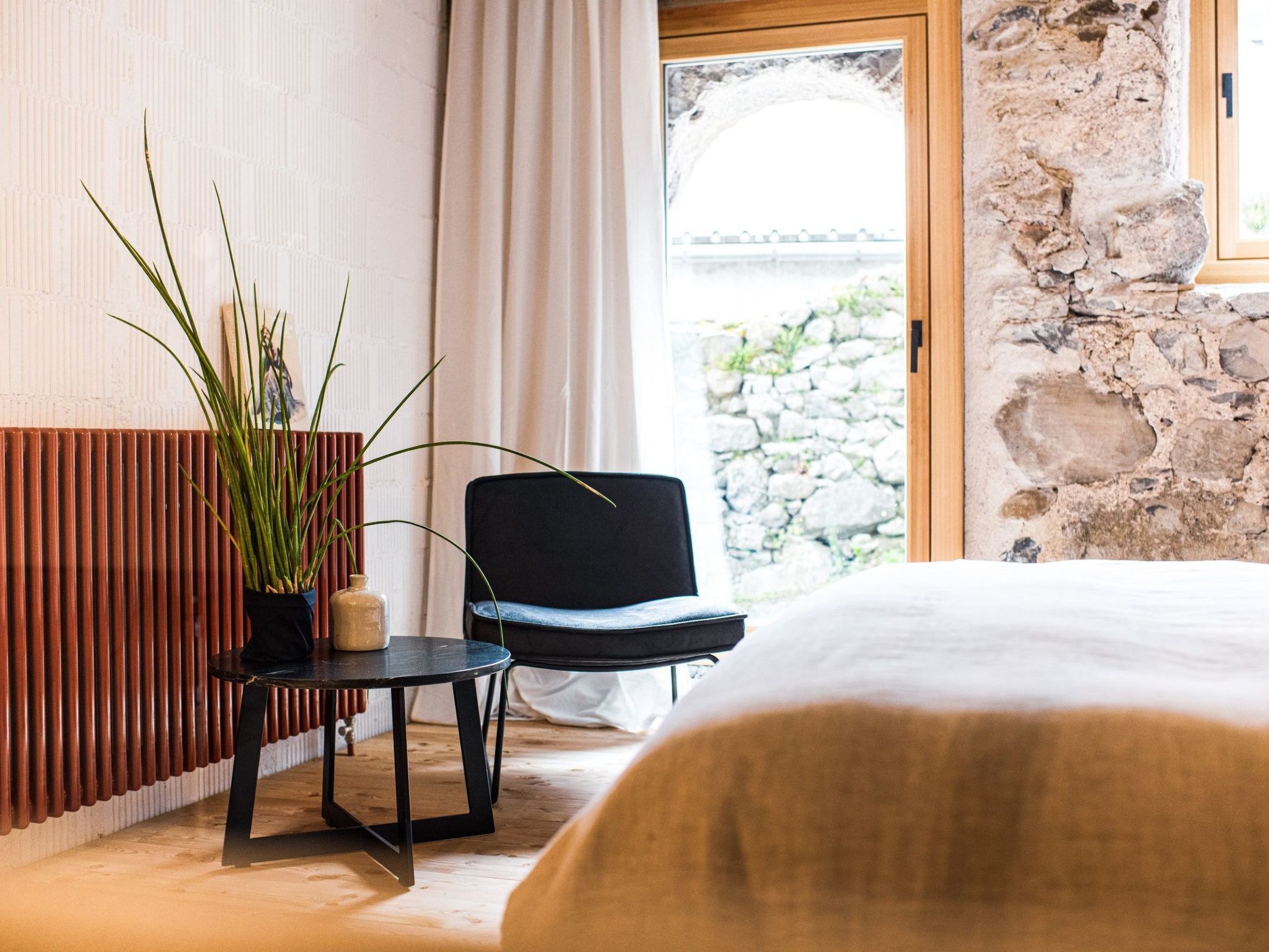 Una habitación con cama, una silla, una mesita, una planta en maceta y una pared de piedra.