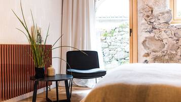 Una habitación con cama, una silla, una mesita, una planta en maceta y una pared de piedra.