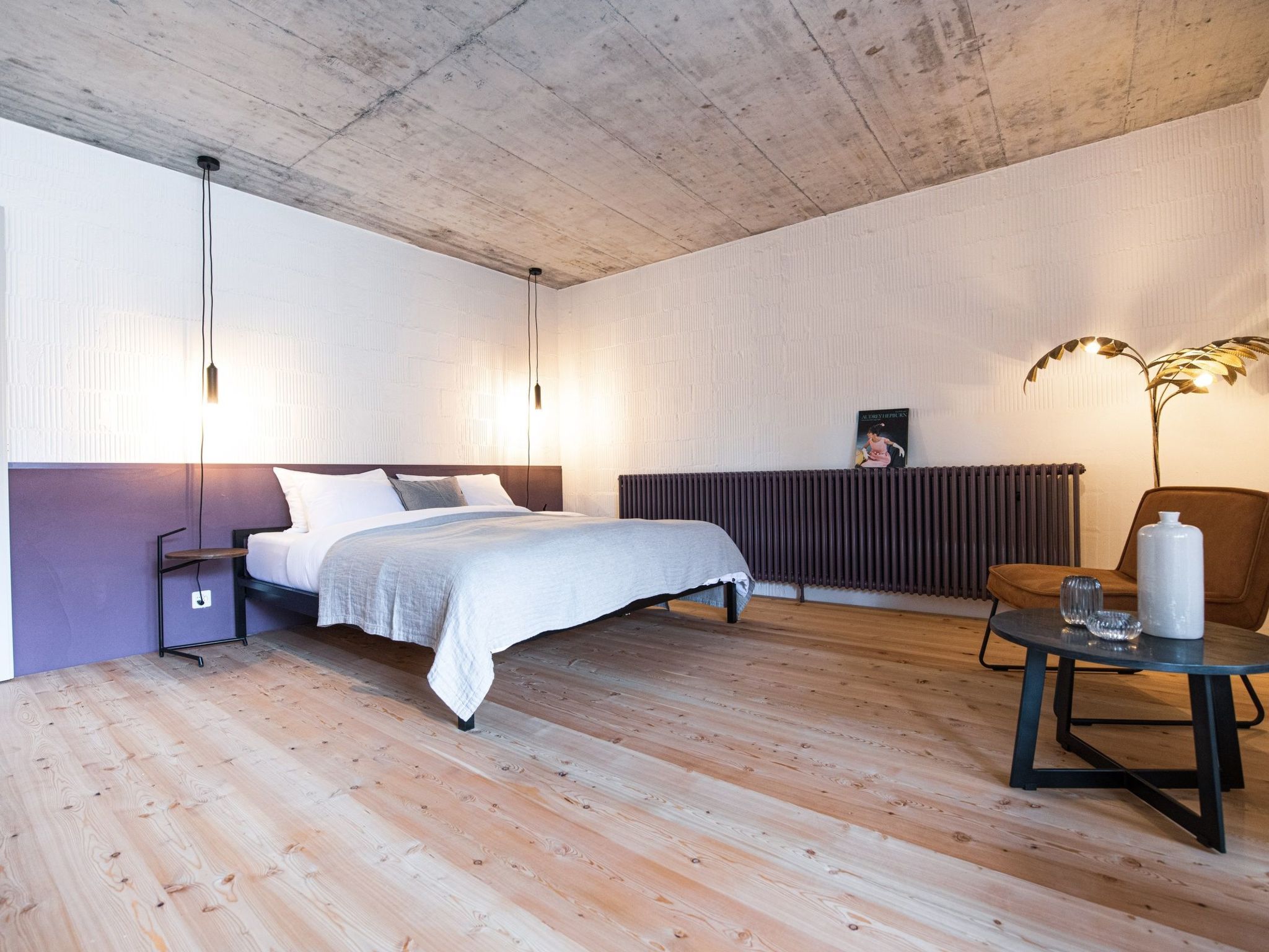 Ein Schlafzimmer mit einem Bett, Nachttischen, einem Heizkörper, einem Sessel, einem Tisch mit einer Vase und einem Wandbild.