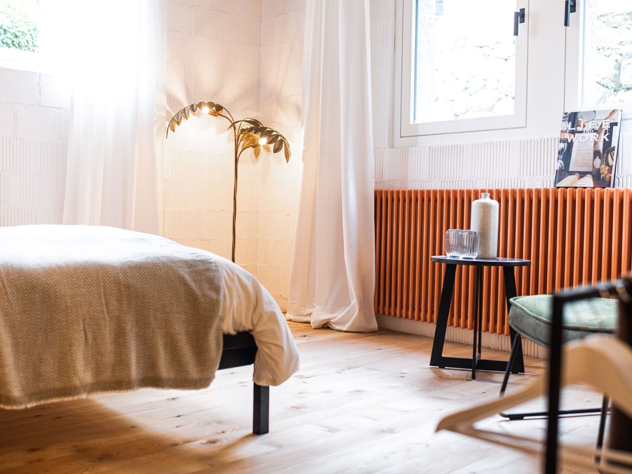 A bedroom with a bed, a floor lamp, a radiator, and a small table with a chair.